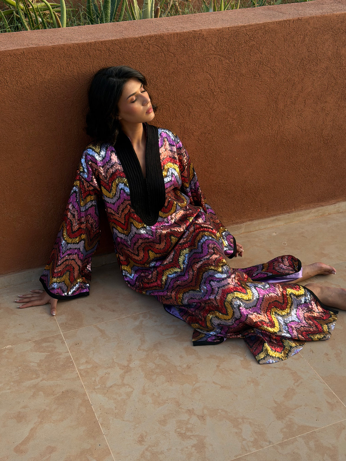 Robe longue à sequins multicolores, modèle assise contre un mur terracotta