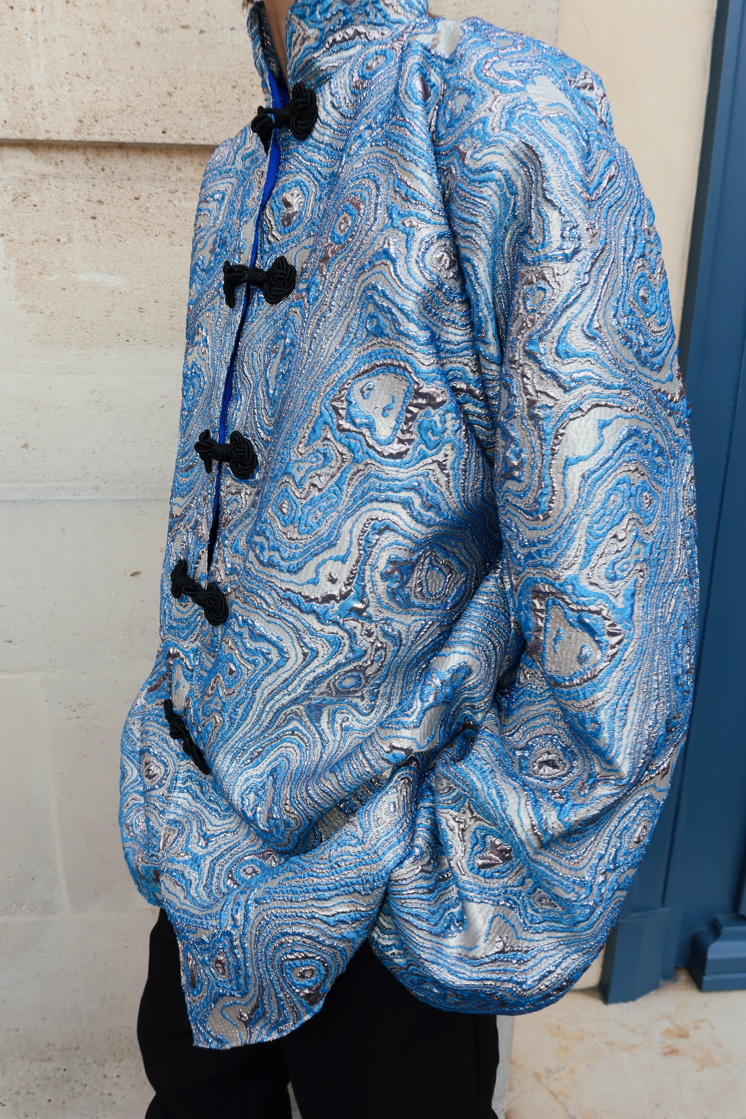Blue brocade jacket seen from the side, highlighting a structured silhouette and black Moroccan hooks